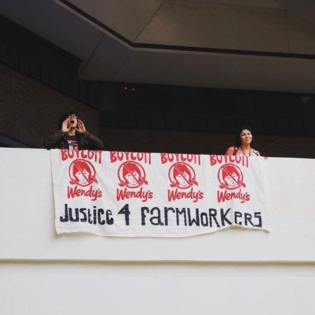 A pixelated, square digital image shows two people on a balcony behind a large banner they've just dropped. One of them is me, on the left, and my head is tilted back as a yell something out. The banner is white and has four red Boycott Wendy's logos printed across it, with the words "Justice 4 farmworkers" hand-painted along the bottom.