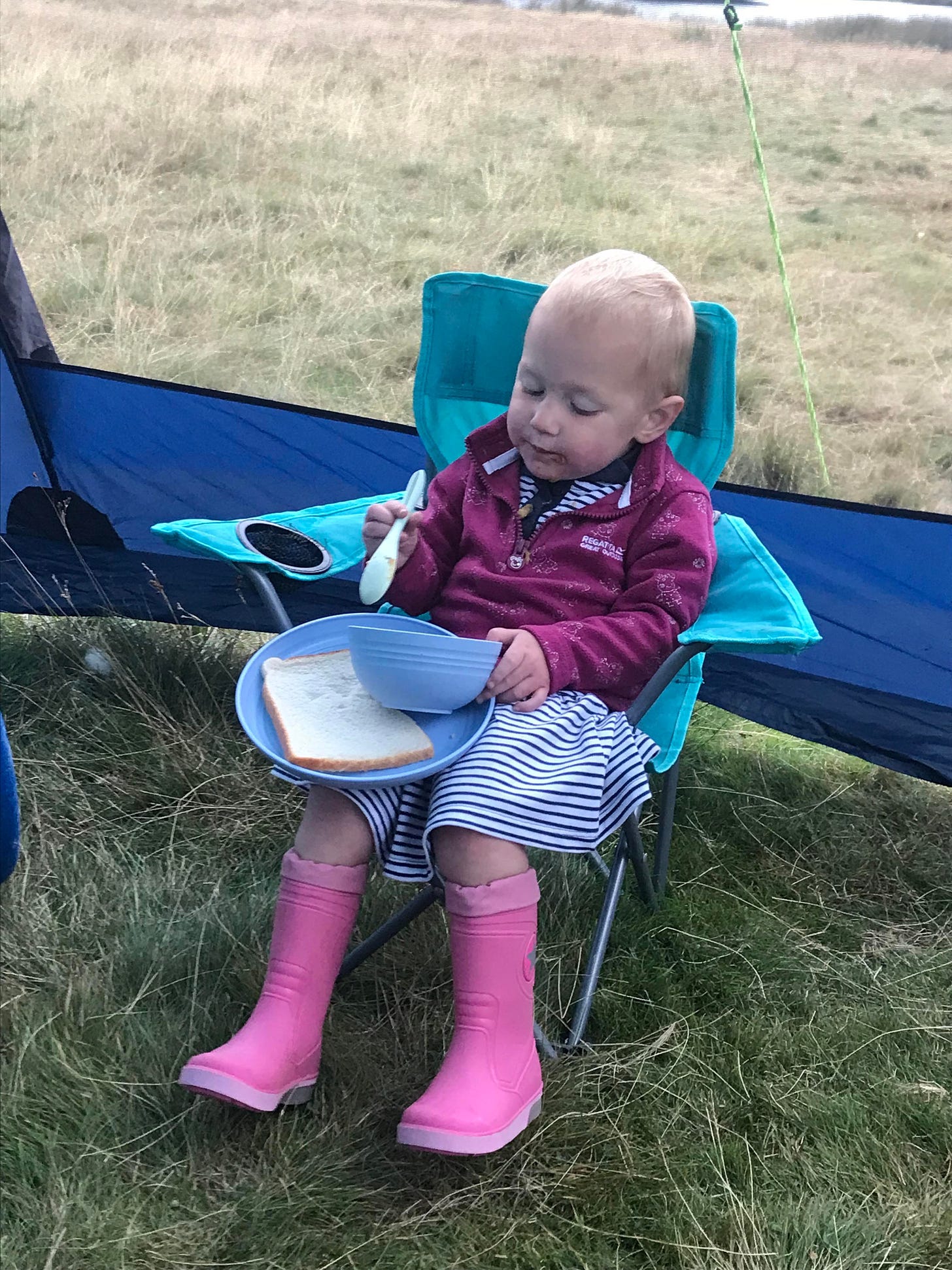 Toddler sat in a camping chair with a plate of bread, wearing pink wellies