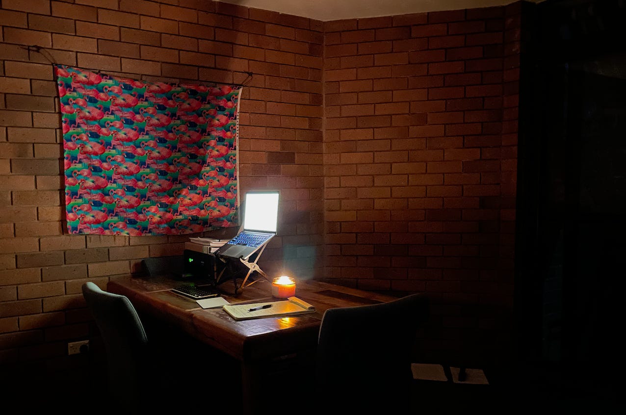 writing desk under flamingo poster with laptop on it, lit candle on desk, brick walls in background, dark lighting all around