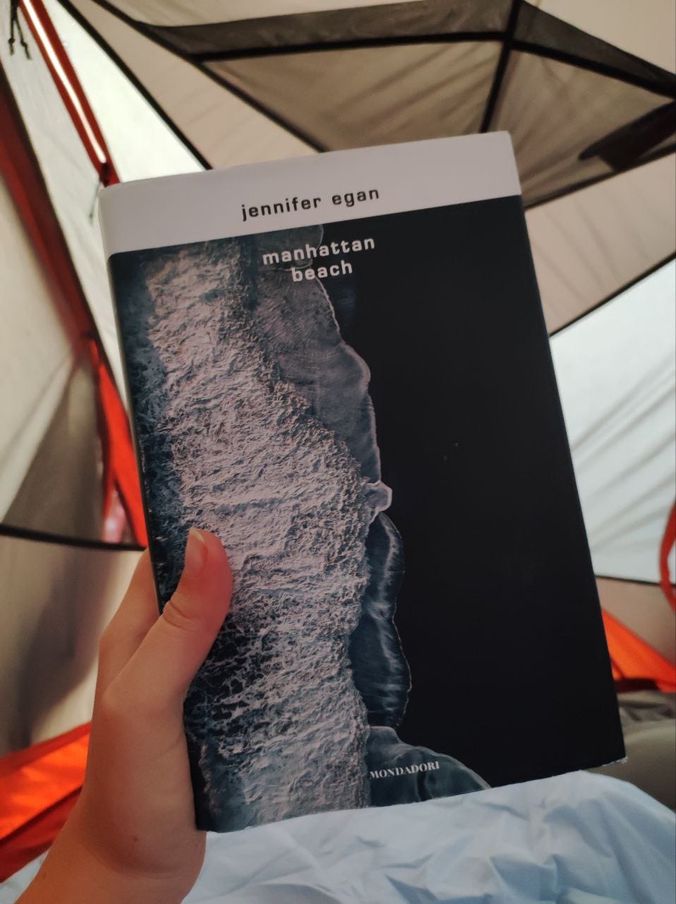 Siamo in una tenda. Una mano tiene un libro. Sulla copertina ci sono delle onde. Il titolo è Manhattan Beach è l'autrice è Jennifer Egan.