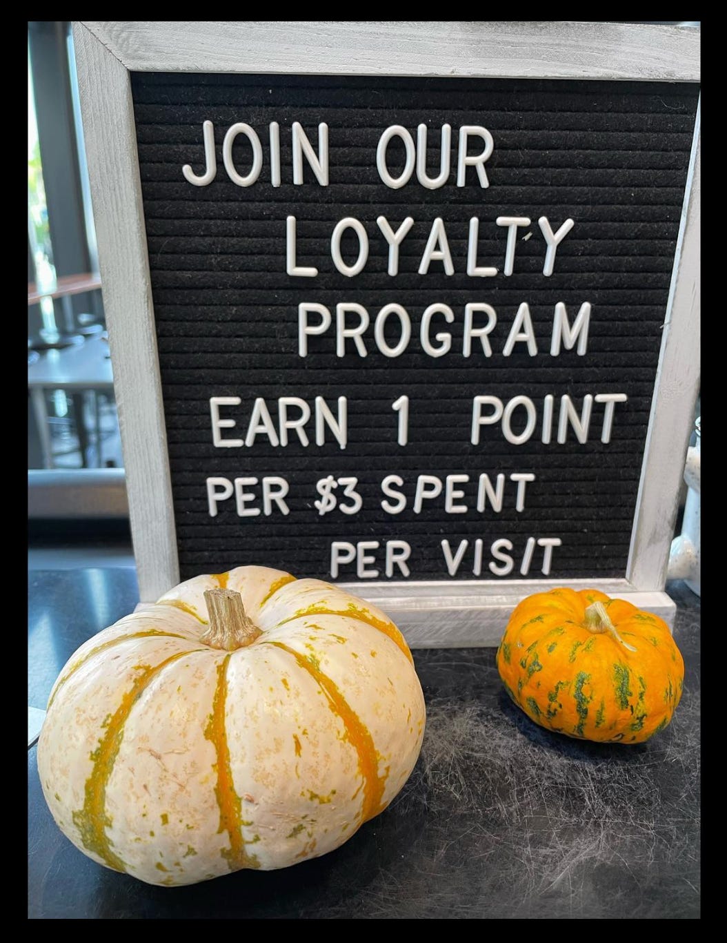 A pegboard menu sign advertising a loyalty program at a coffee shop. Two decorative pumpkins prop up the sign.
