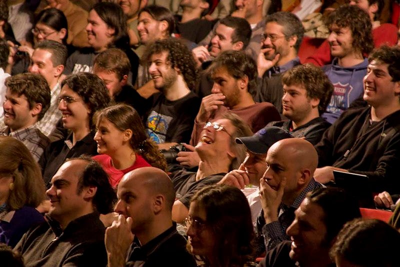 An audience of mostly white men and maybe two white and one Asian women, laughing at some jokes. 