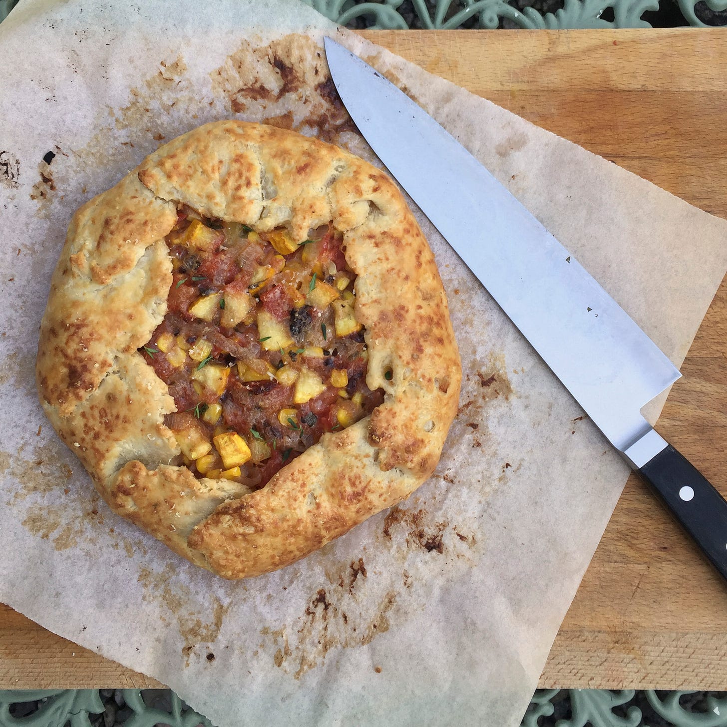 A browned galette filled with tomatoes, corn, and yellow zucchini, sprinkled with thyme. It sits on a piece of parchment, browned from the oven, atop a wooden cutting board. A large chef's knife sits next to it at an angle.
