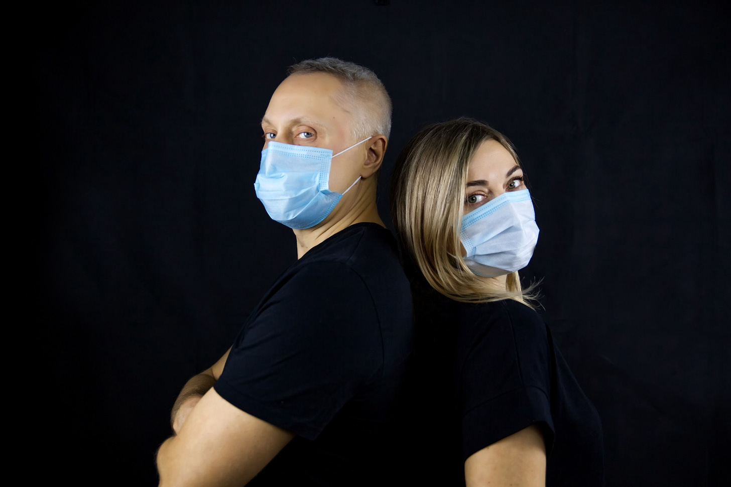 A serious couple in blue face masks stand back-to-back against a black background.