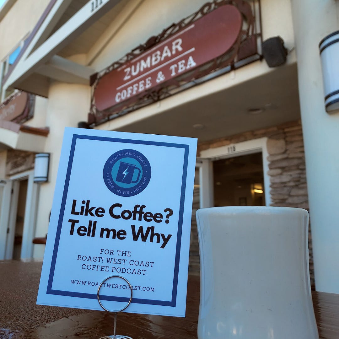 A close-up on a white coffee mug next to a paper sign that says "Like Coffee? Tell me why." The front door and sign for Zumbar Coffee and Tea is in the background.