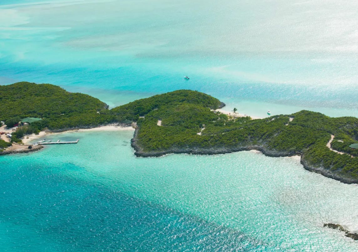 A tropical island surrounded by aquamarine water. On the far left of the island land visible is a complex of buildings set very close to a private beach. There is a small wooden pier with two little boats moored off them. The rest of the land is covered by trees and greenery.