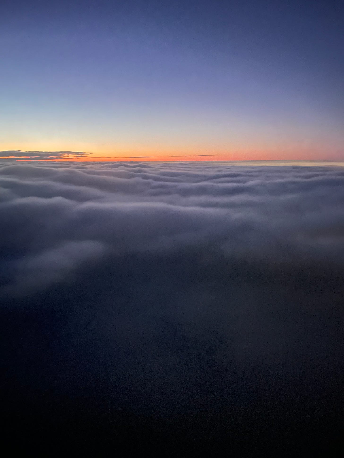 Above the clouds, the dawn chases the darkness. Bright orange begins to fill the horizon, creating a orangish-dark blue gradient above the clouds.