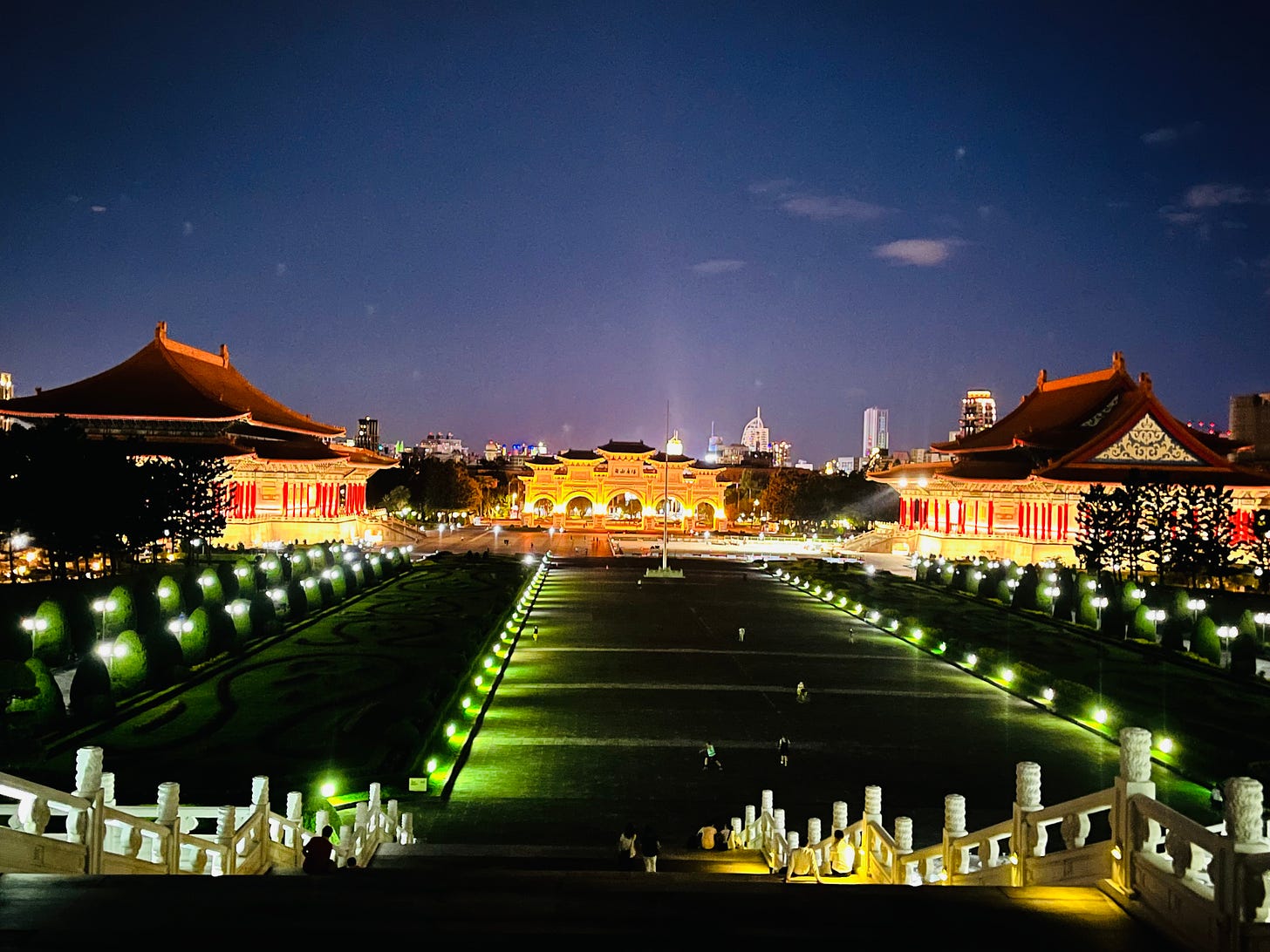 Two almost-identical performance halls, built in the style of Chinese palaces, flank a dark boulevard that ends at a grand white archway with upturned eaves. Everything is brightly illuminated for nighttime visitors.
