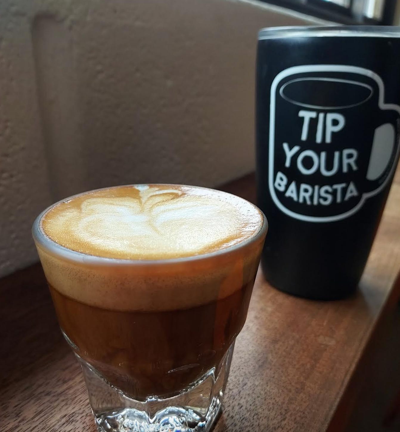 A close up of a cortado-espresso topped with steamed milk-in a rocks glass in front of a white concrete wall and sitting on a wooden countertop. A black tumblr with a Tip Your Barista sticker on the side is blurred in the background.
