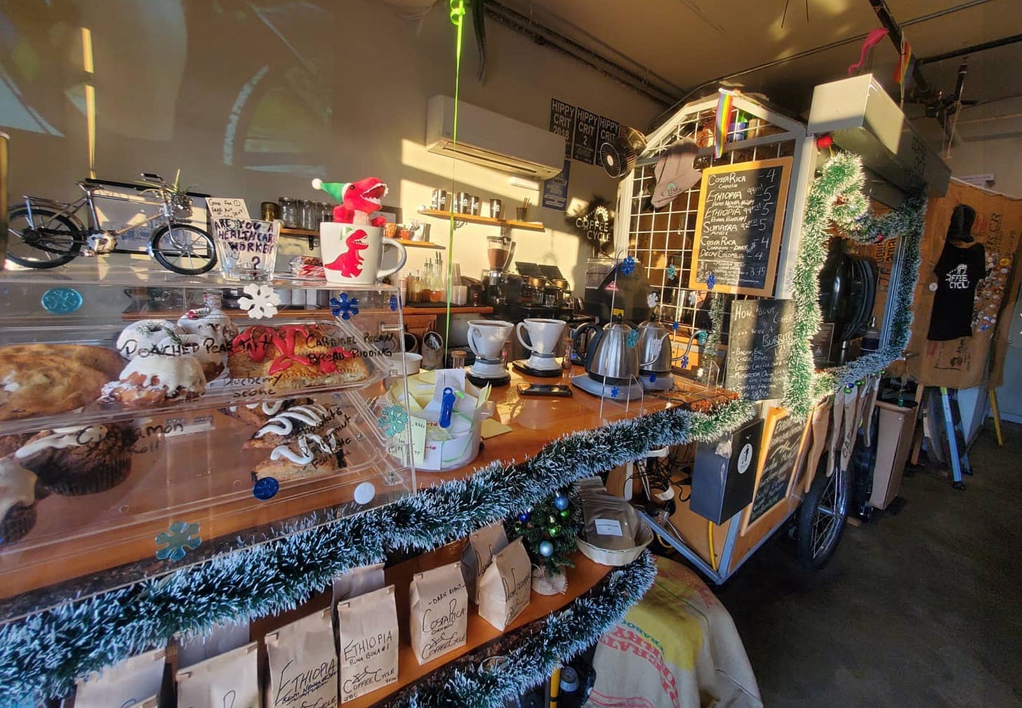 A long shot from left to right of the coffee bar at Coffee Cycle cafe in Pacific Beach San Diego. Wood bar top with coffee kettles, a pastry card, a dinosaur christmas decoration and on a shelf below brown bags of coffee beans for sale with handwritten labels.