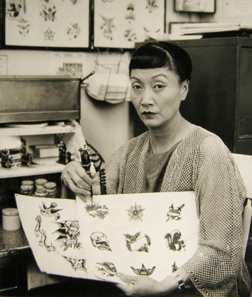 an older Anna May Wong sits in a tattoo parlor and points to a tattoo design with her needle
