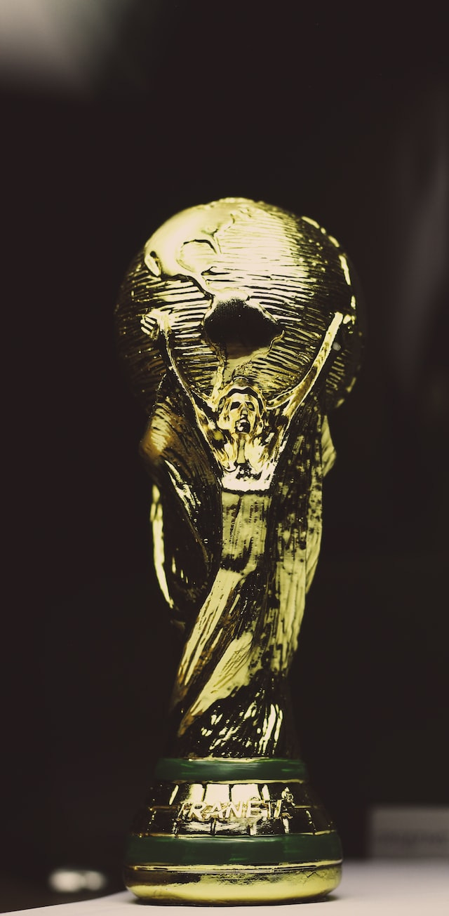 The FIFA World Cup Trophy, a golden statue of a woman whose aloft arms are holding behind her a globe, with the Americas visible in the front. The trophy sits at the edge of a table, against a black background. Light is coming down from the top left corner.