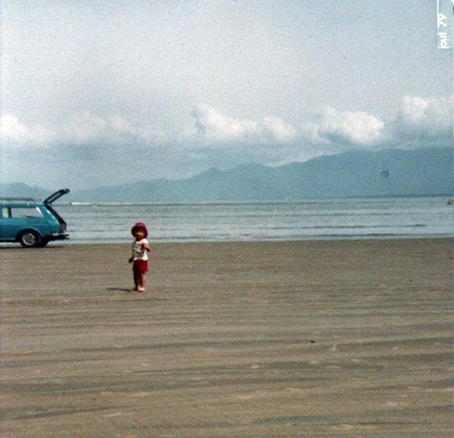 foto de uma criança bem pequena, de chapéu vermelho, na areia de uma praia, com uma variant azul atrás, o mar e algumas montanhas