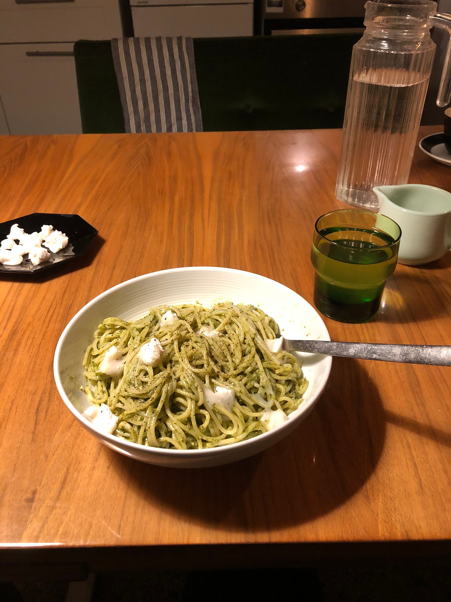 A wide shallow bowl of pasta with a green pesto, with dotted white cheese over the top. A fork lies into the bowl of food. The bowl is on a warm wooden table, toplit. Further back on the table, to the left, is an octagonal black dish with more pieces of cheese. To the right, a cast clear glass jug of water, a green glass of water and a china jug. 