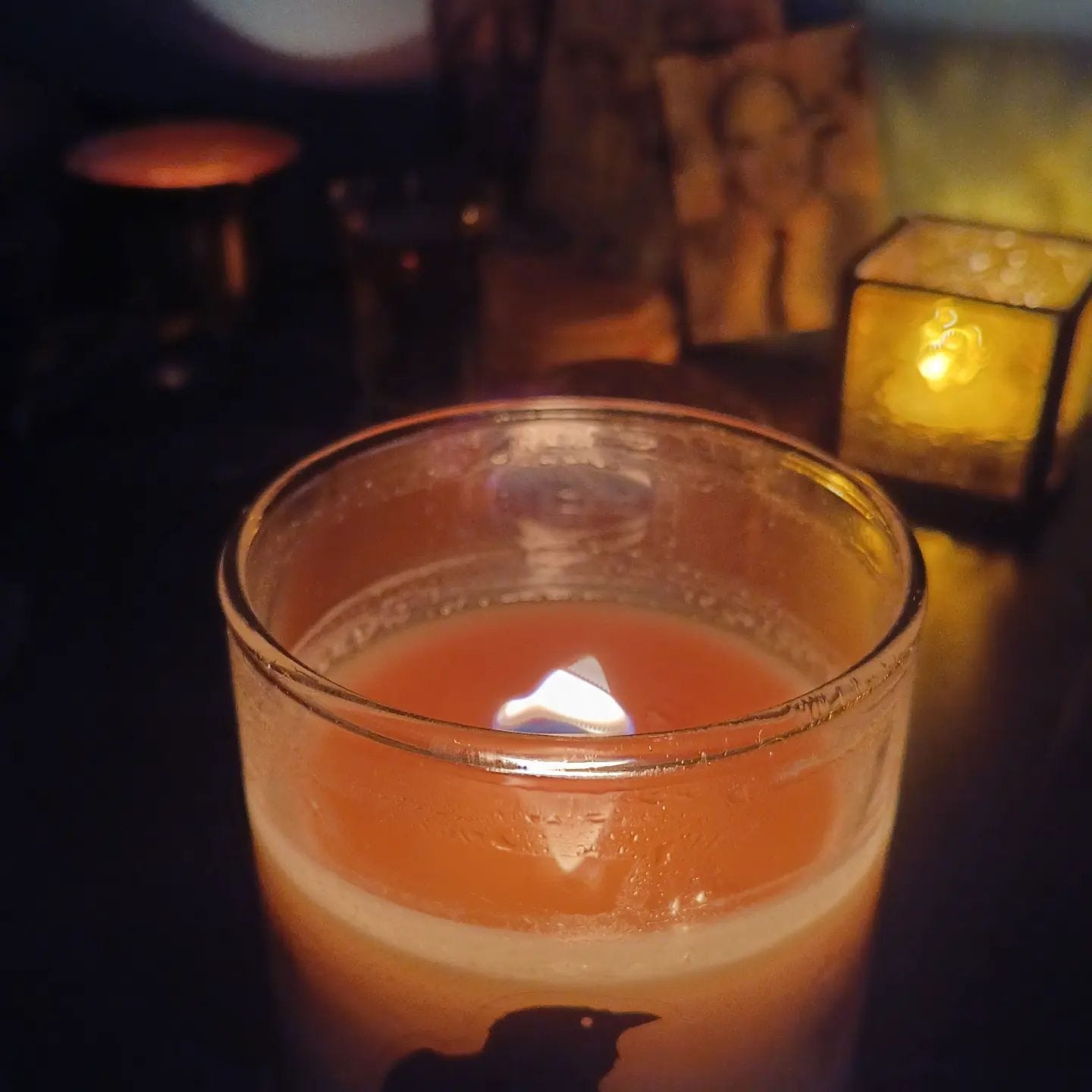 a lit candle with a black raven on the jar, over a background of my ancestor shrine in shadow