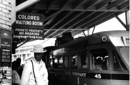 Photo at a railroad station with a sign that reads "colored waiting room"