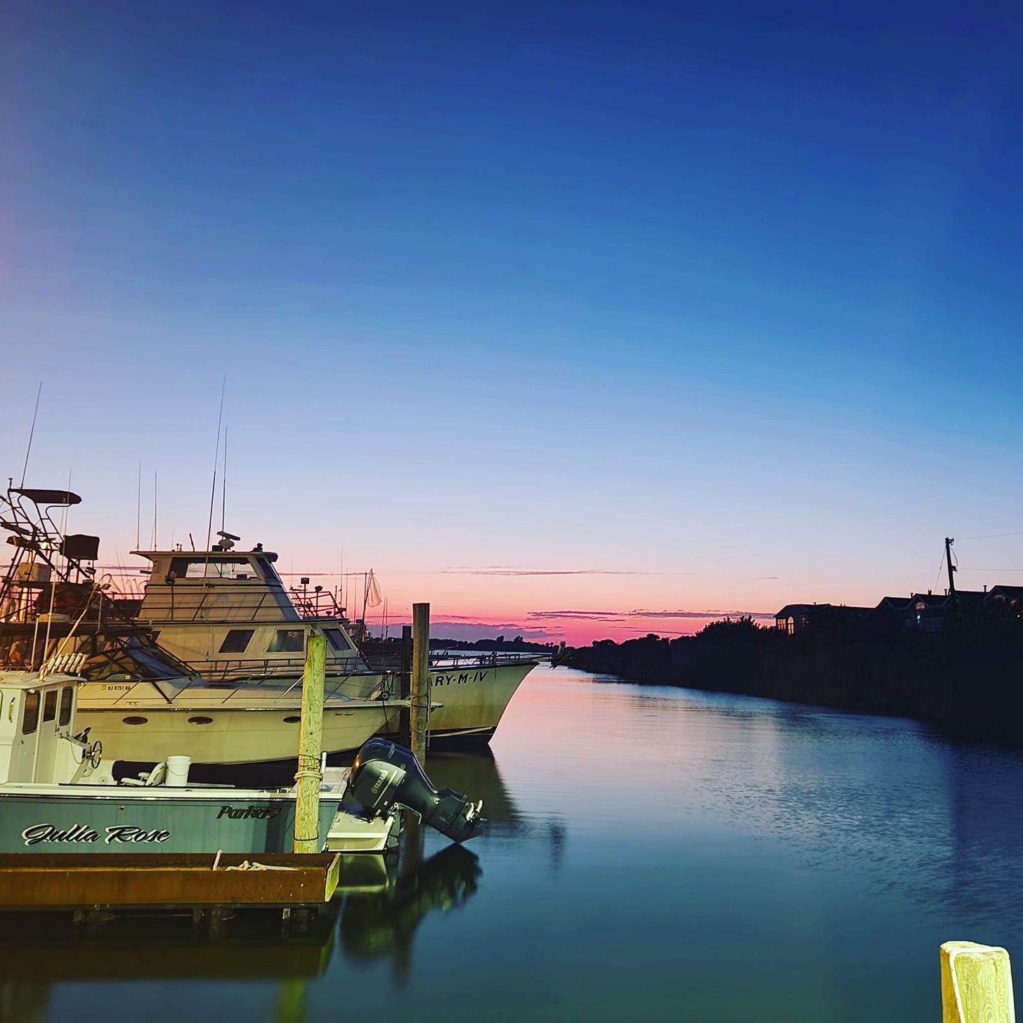 A rich blue sky and a pink line of sunset behind several dirty fishing boats in a blue harbor.