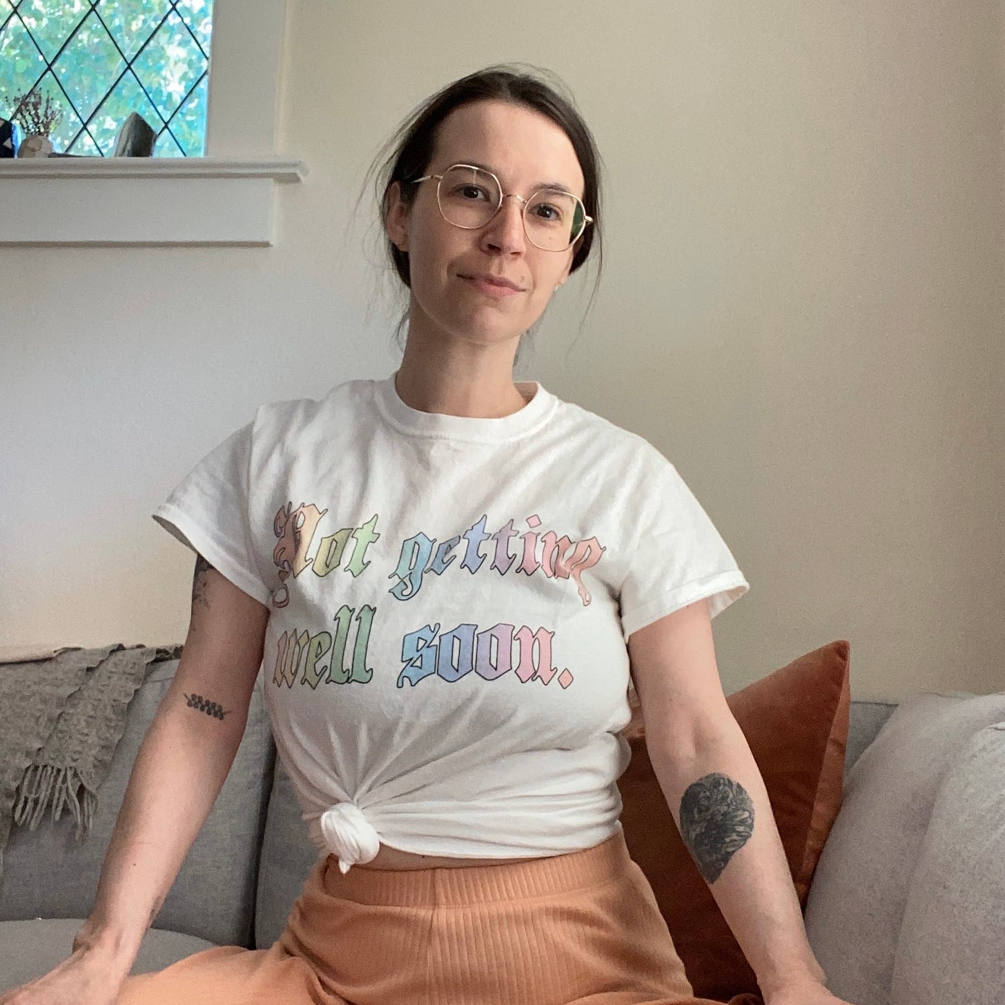 Image of a 30 something white woman with pulled back brown hair and wife frame glasses, sitting up wearing a white t-shirt that reads “not getting well soon”