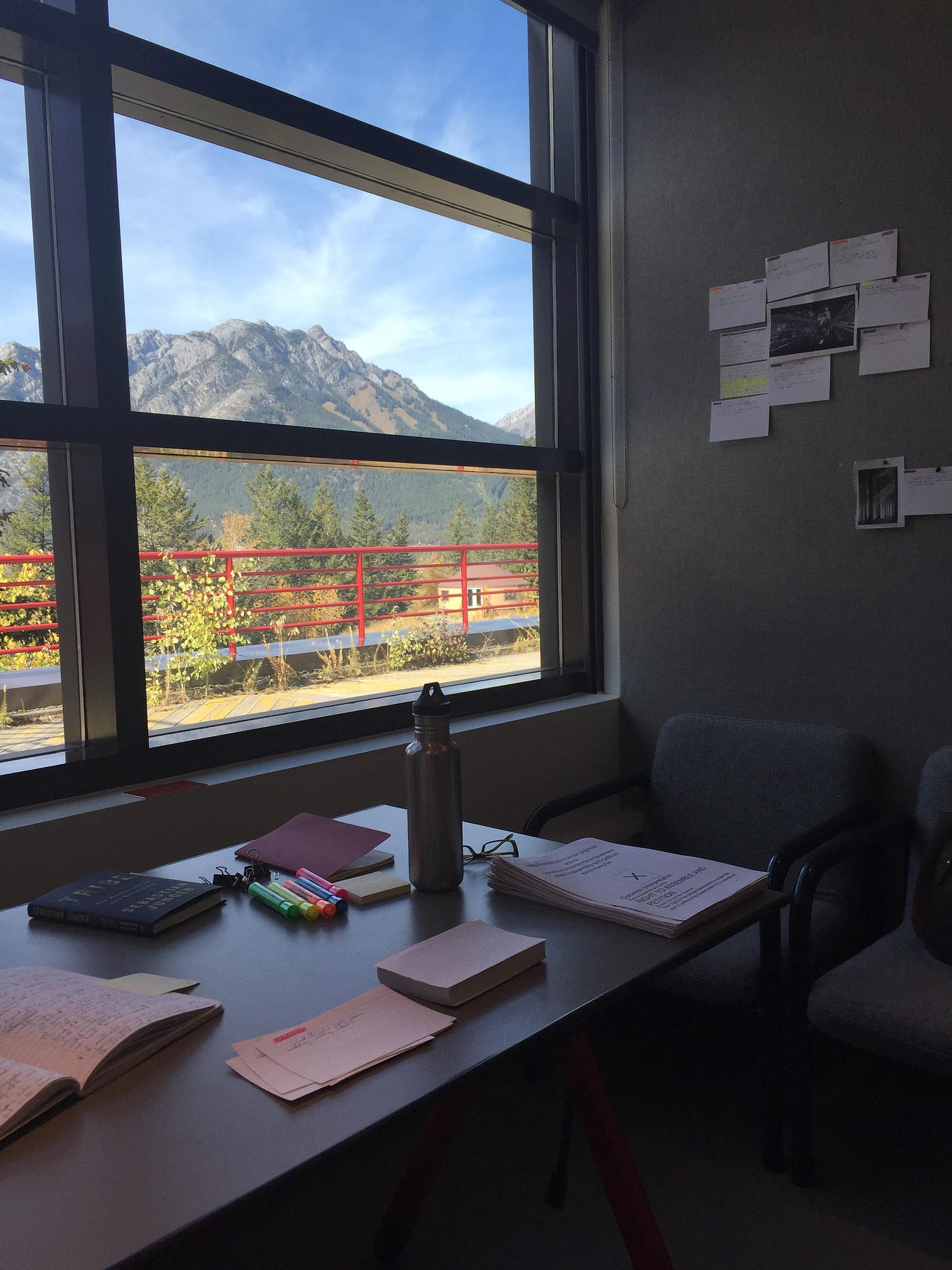 A desk with papers and highlighters, index cards, and books, in front of a window looking out toward the Canadian Rockies