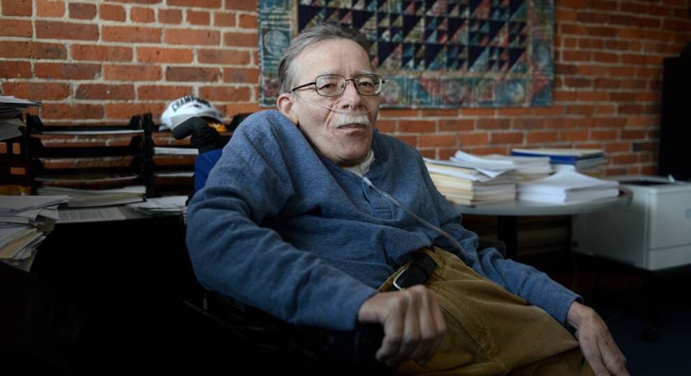 A man in a wheelchair, wearing a blue sweater and tan corduroys sits in an office against a brick wall. Books lay on the desk behind him.