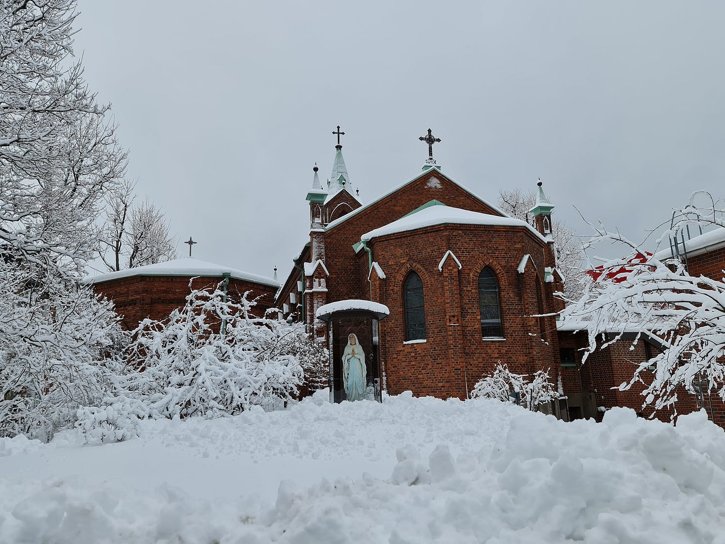 En kyrka i rödtegel med en Mariastaty framför kyrkbyggnaden. Runt kyrkan och statyn är det stora mängder snö.