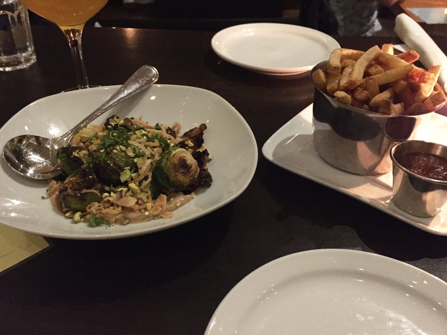 two dishes in the centre of a dark table, one with brussels sprouts covered in crisp fried shallots and nuts, the other a plate holding a metal cup of fries and a small metal ramekin of ketchup. Two side plates are visible in the background and foreground.