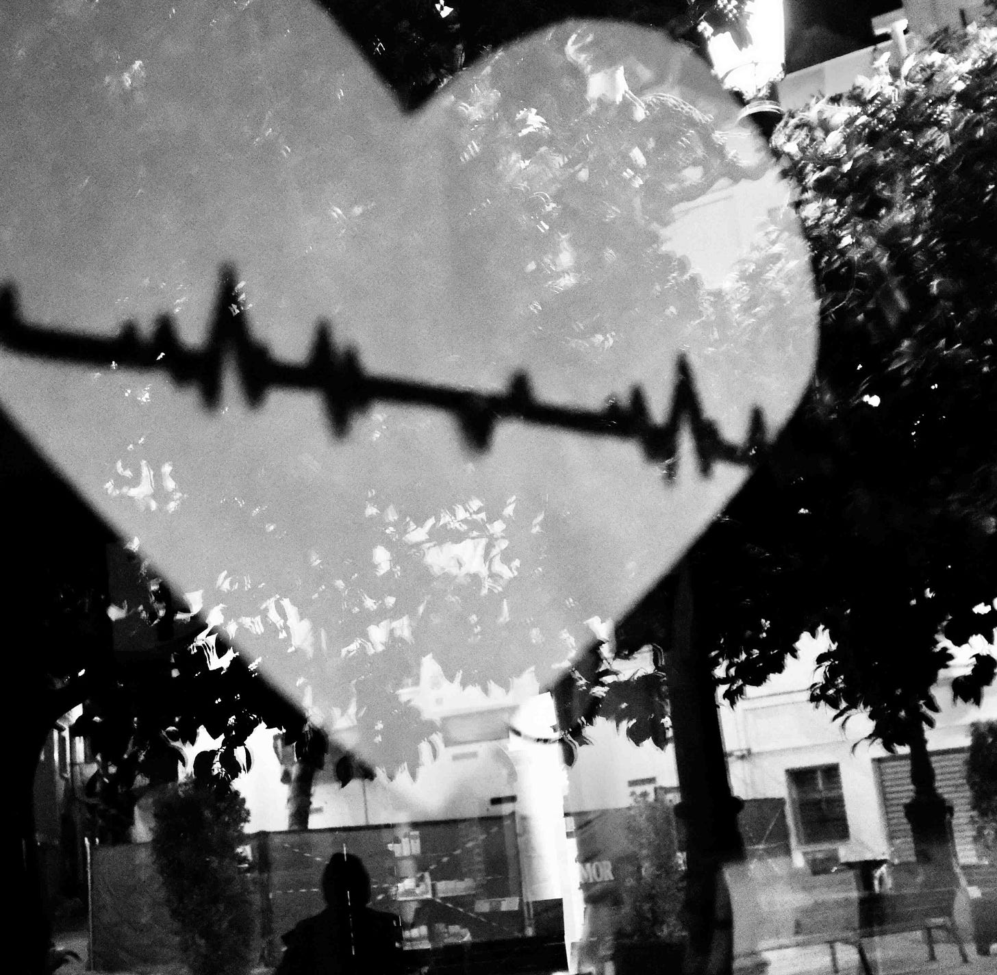 A black and white image of a large heart with a jagged line running through it. It is on a window which reflects a street scene and trees.