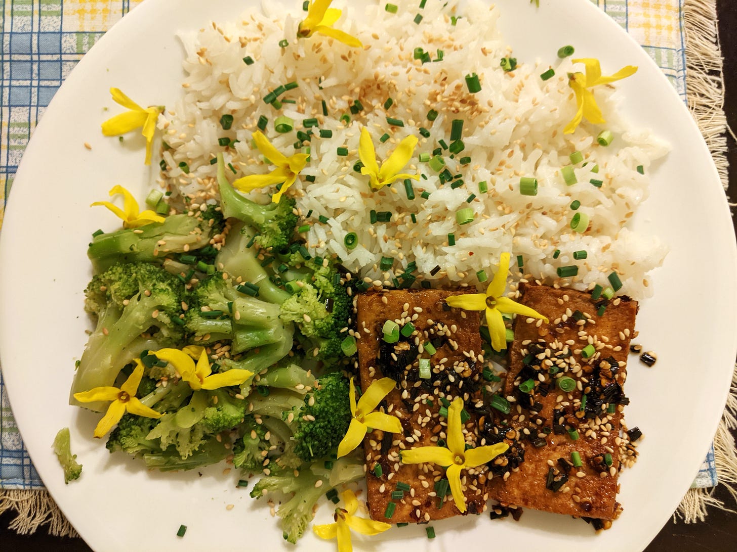 A plate with rice, broccoli, and tofu garnished with sesame seeds, field garlic, and flowers.