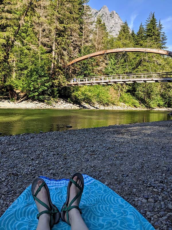 bridge arching over the Middle Fork Snoqualmie River beneath Stegosaurus Butte, from a beach towel view on a gravel bar on the banks of the quiet green river