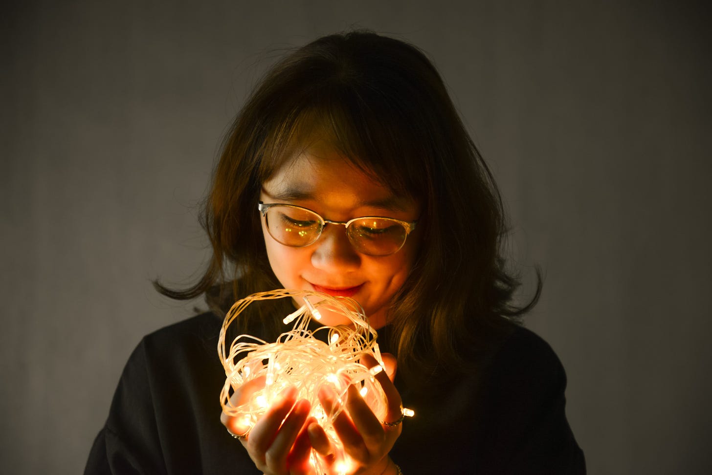 a young girl, wearing glasses, cups in her hands fairy lights. She has a soft smile on her face.