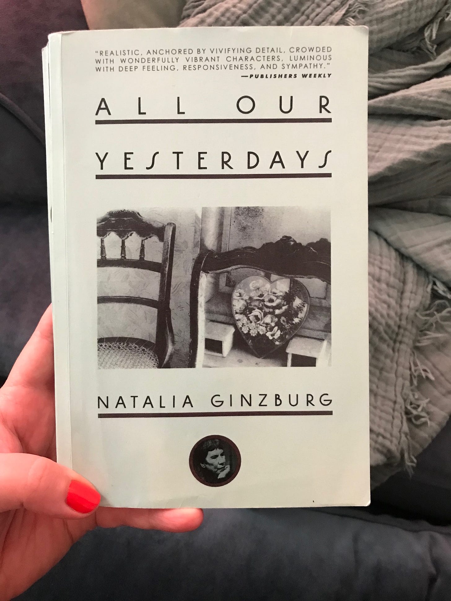 Hand holding a book called All Our Yesterdays by Natalia Ginzburg, with blue blanket and chair in background