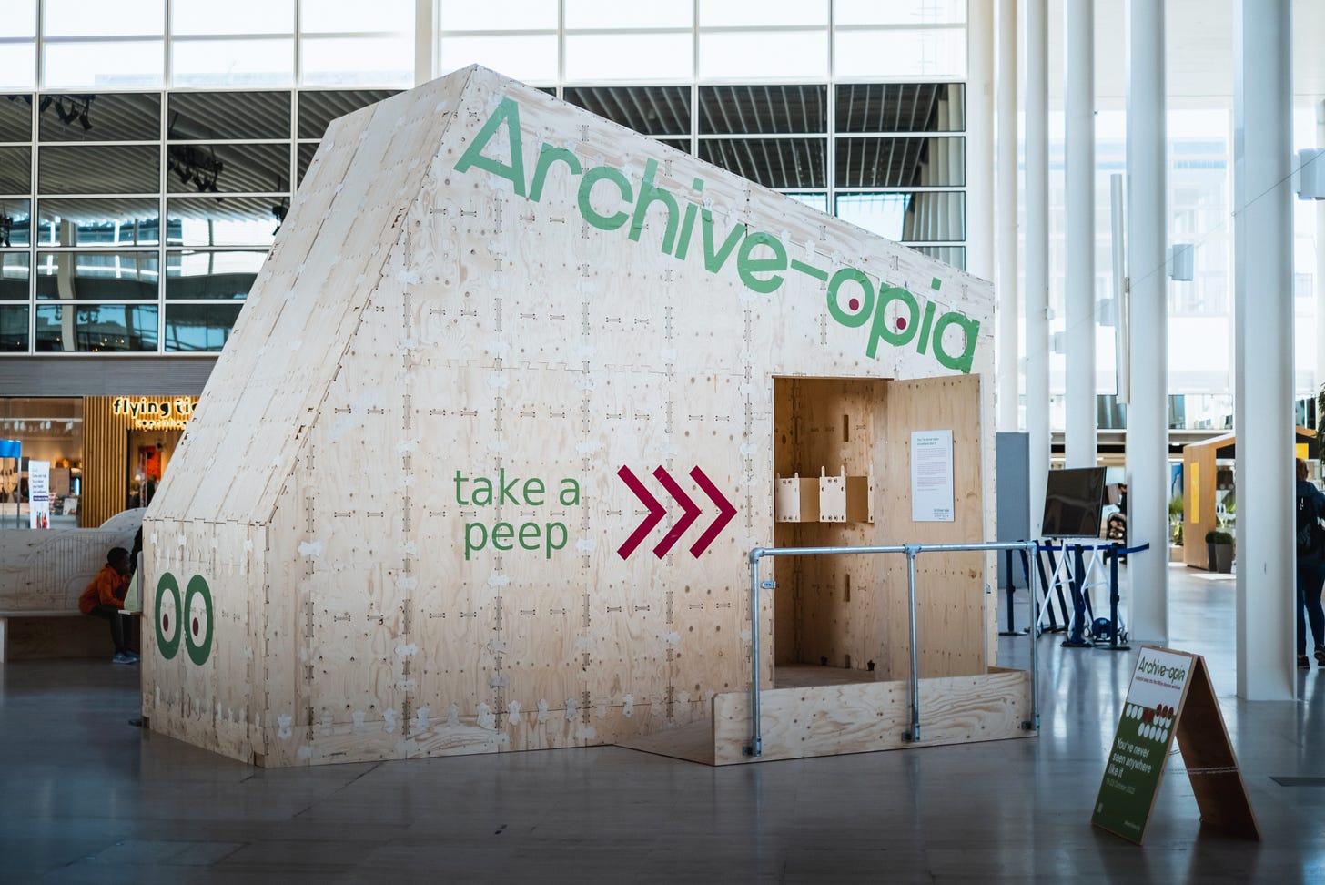 An image of the finished WikiPavilion project. The structure sits inside a shopping mall and has the words "Archive-opia" "take a peep" printed on its front