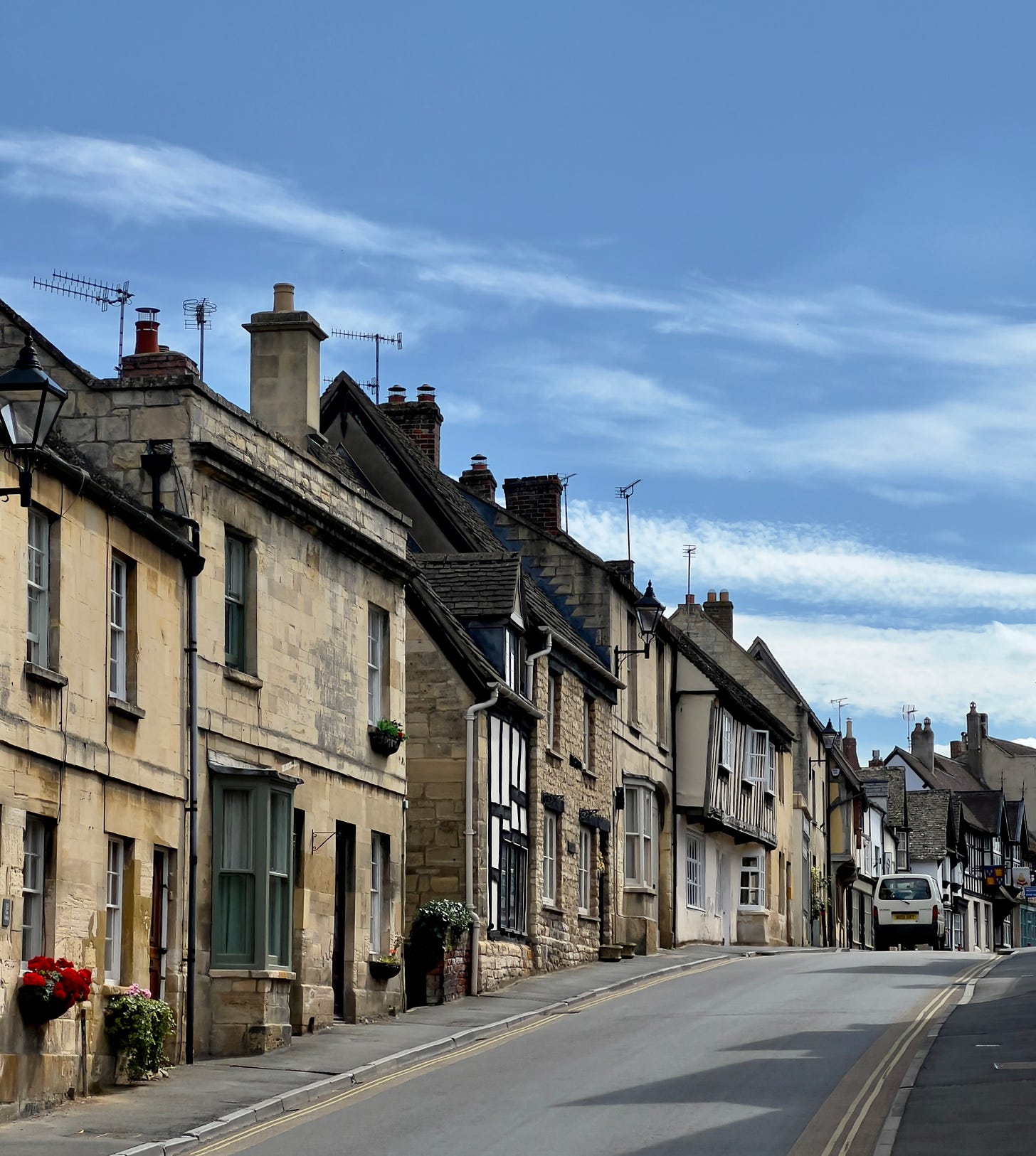 Street scene from Winchcombe, England with yellow stone buildings, red geraniums  in a window box and a blue sky with white clouds