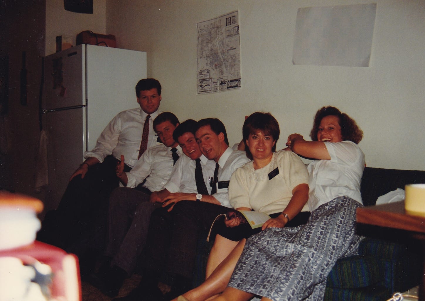 Six Mormon missionaries (four male and two female) are crammed in a row onto a small couch in a messy apartment with a map on the wall behind them.