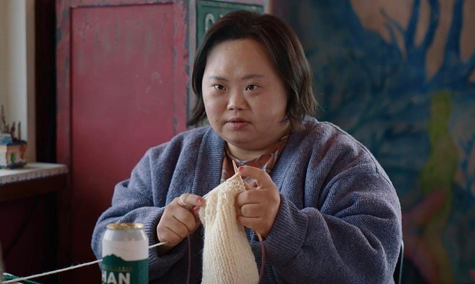 A young woman with Down Syndrome sits by a window and knits.