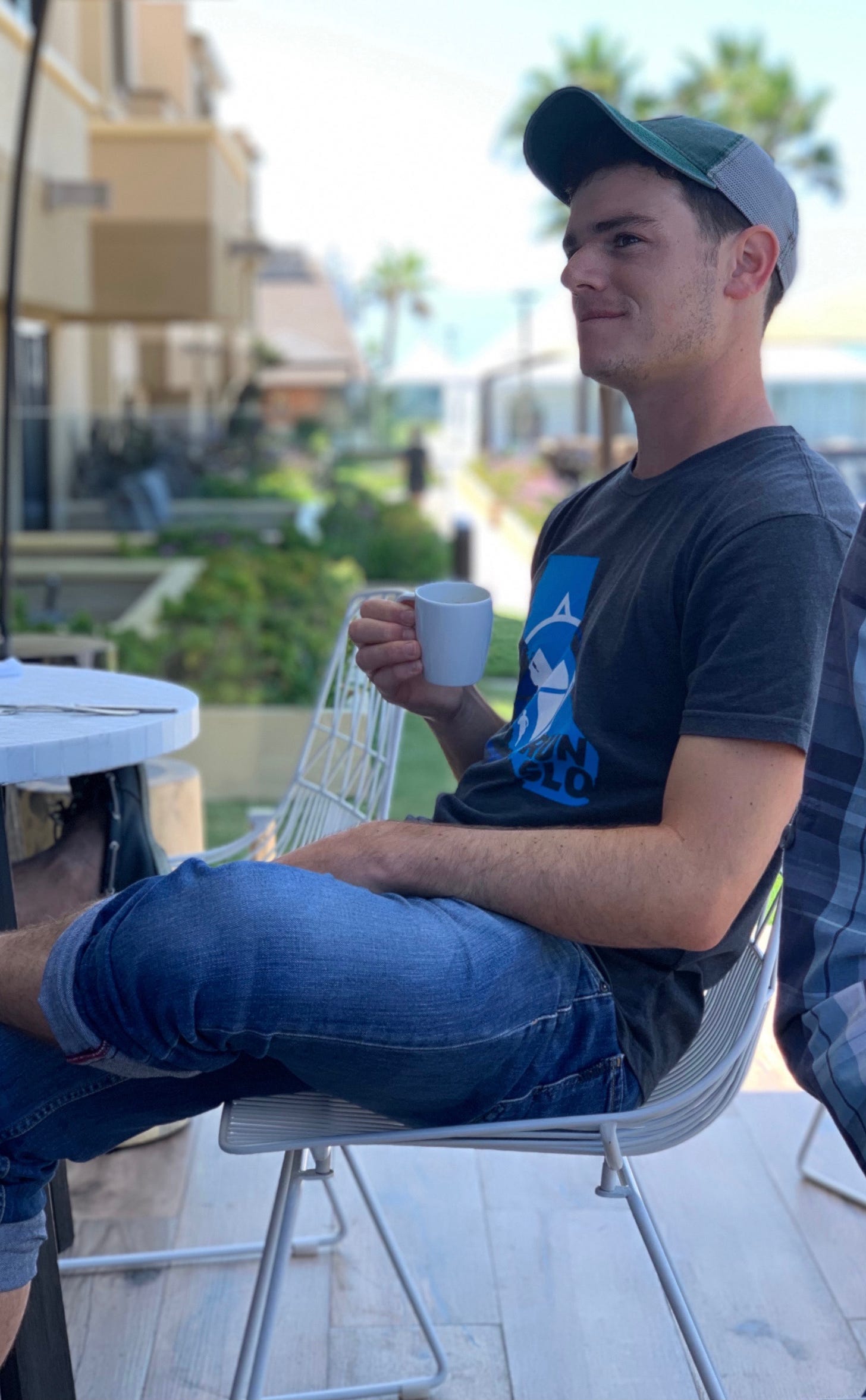 Profile photo of Brendan Abrams, a white man in a blue t-shirt, jeans and baseball cap sitting on a patio chair holding a cup of coffee while looking off into the distance. A palm tree and blue water next to a walking path is in the background.