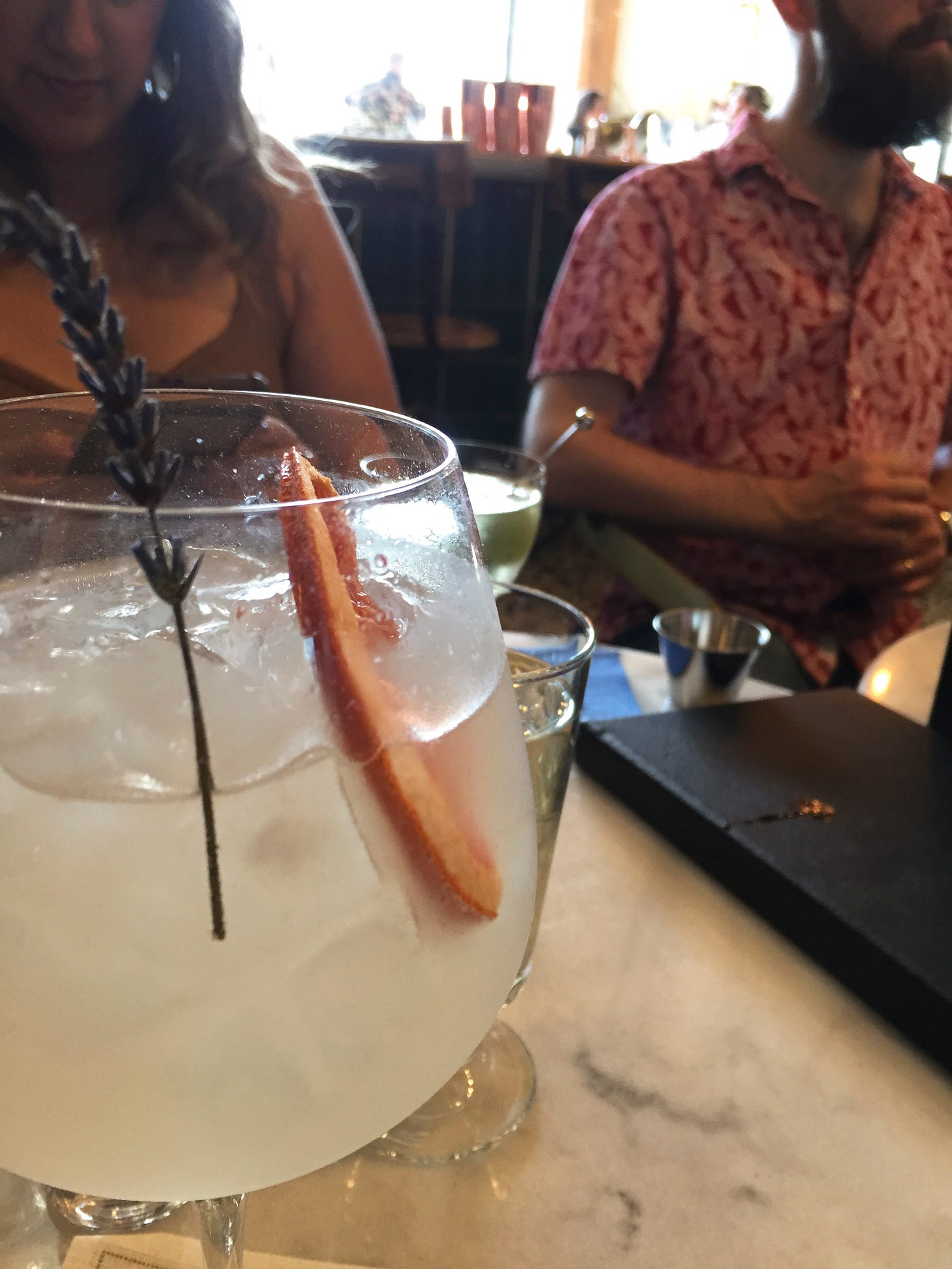 A close up view of a wine glass with a pale cocktail garnished with a sprig of lavender and a slice of dried grapefruit. In the background across the table are two people each with cocktails of their own.