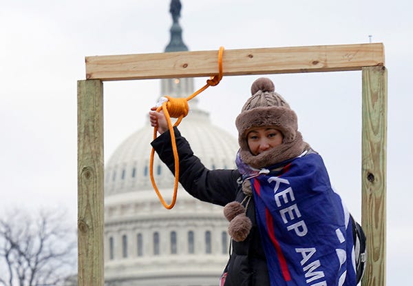 A woman at the insurrectionist riots at the Capitol brandishes the noose in the makeshift gallows set up by the supremacists who came on January 6 to overthrow democracy and install a dictator. She wears a Trump flag around her shoulders. The Capitol Building can be seen between the gallows uprights.