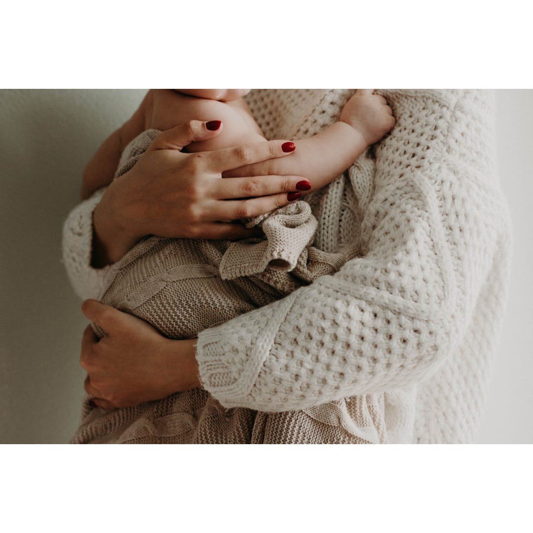 Photo of a white mother holding her baby tightly to her chest. The image is cropped such that their faces do not show. The mother is wearing a white sweater and the baby is wrapped in a pale beige shawl, but the mother's nails are painted red.