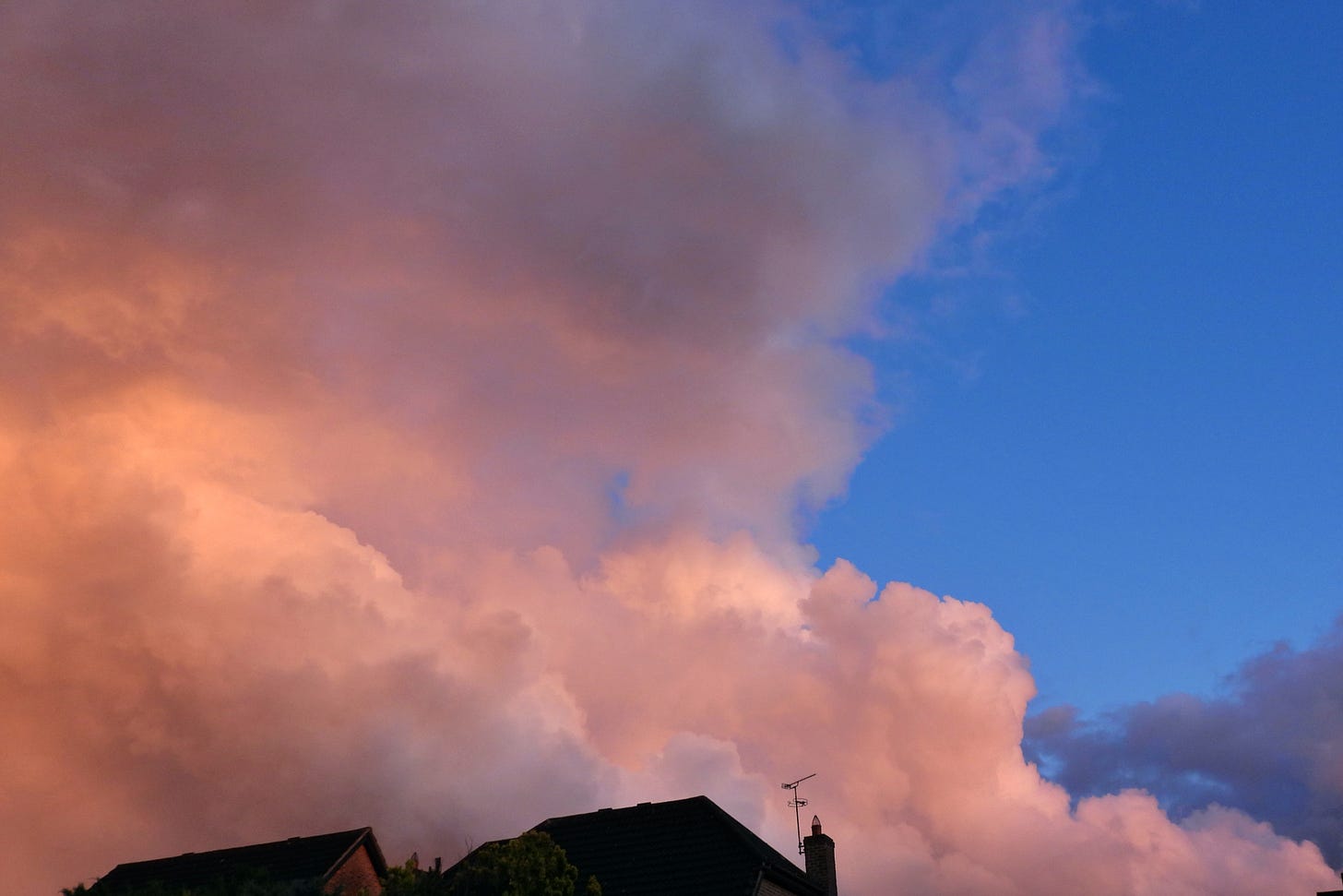 Looking up at the sky showing cumulonimbus clouds against a blue sky. The cloud is glowing with a peachy light from the sunset. House roofs are visible in the bottom of the image.