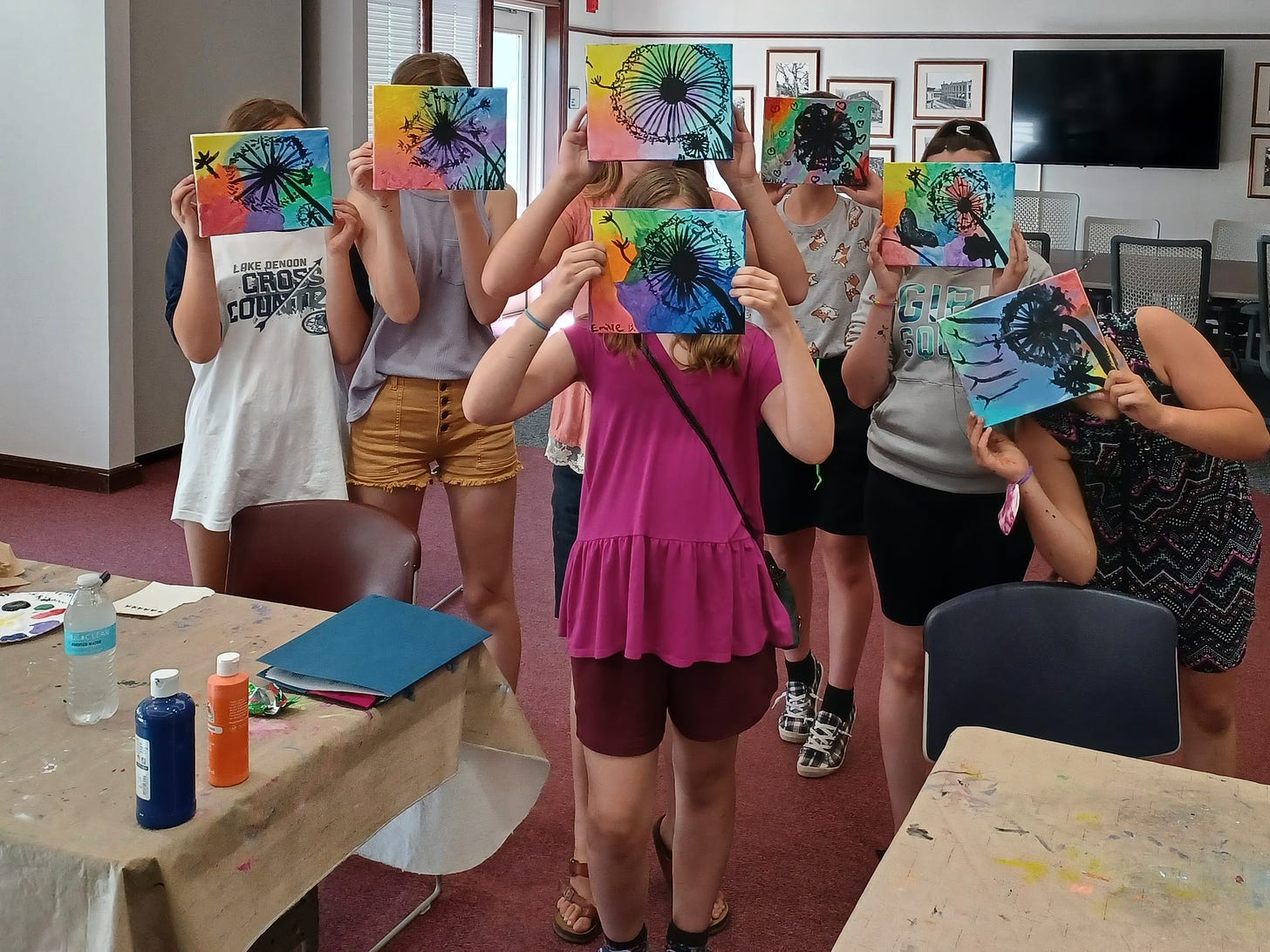 A group of young people hold colorful artworks over their faces in a library makerspace.