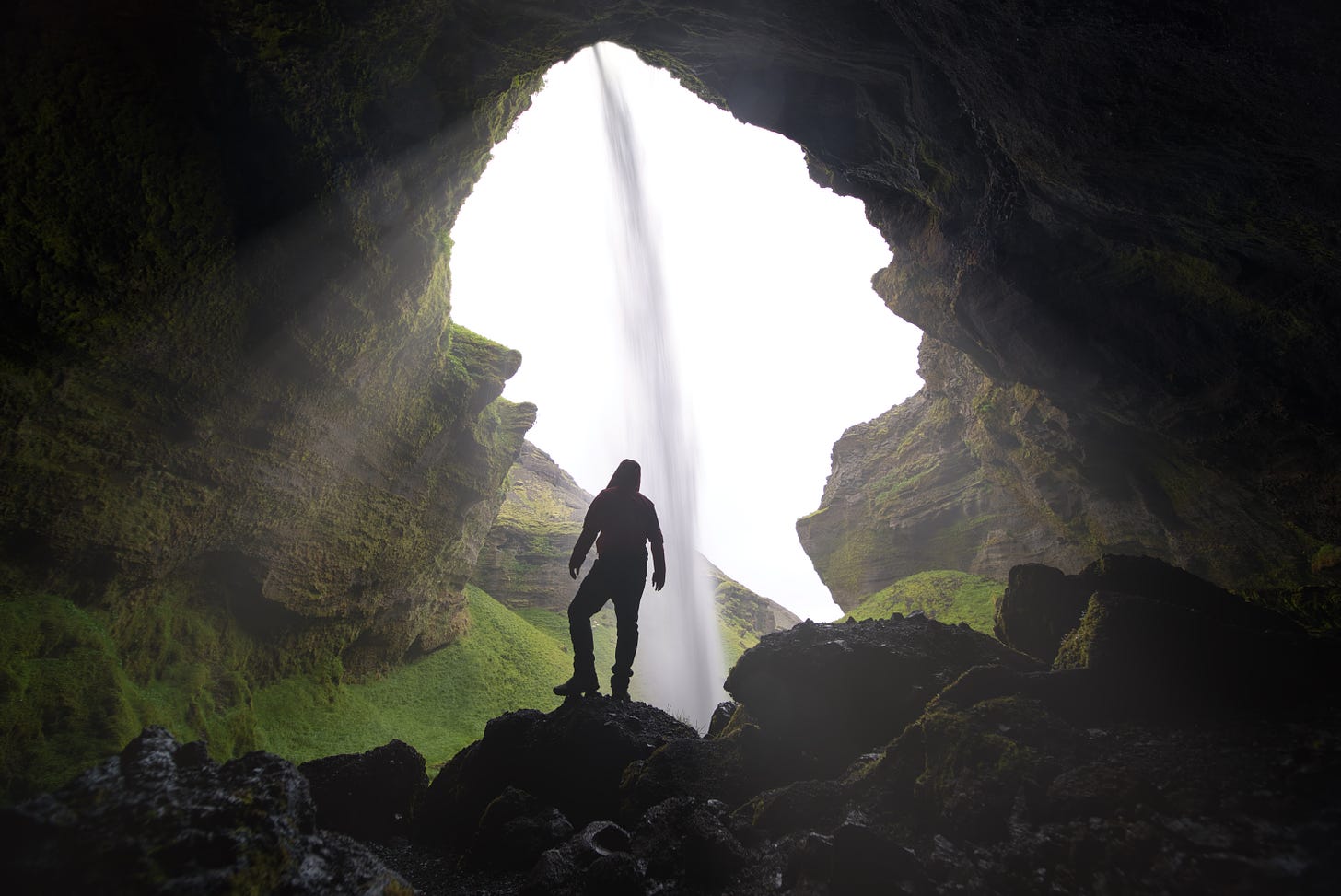 Person standing on rocks at the mouth of a massive cave with a waterfall falling into it from the outside