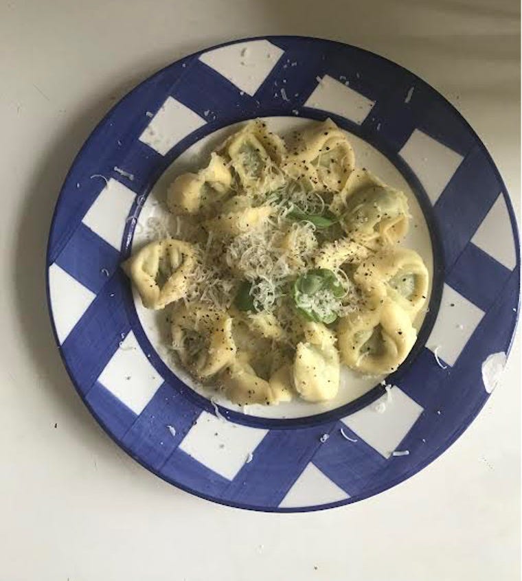 A plate of pasta from above. The plate is painted with thick strokes of blue, criss crossing. On it, tortellini rounds dusted with grated cheese and a few basic leaves. 