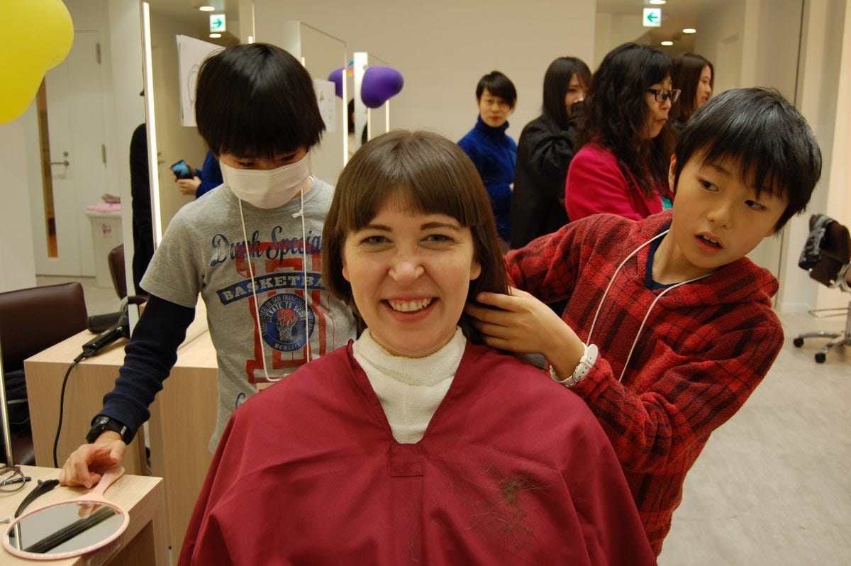 A middle aged woman sits in a salon chair, caped and ready for a haircut, smiling, while two young boys, age around 10, stand behind her, prepping to cut her hair. Other adults in the salon look on in the background.