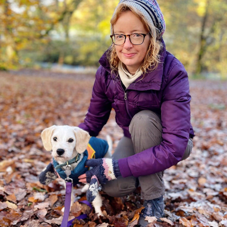 a white woman wearing glasses is smiling as she crouches down next to a super cute dog with short, wiry hair. They are in a woodland, with autumnal leaves strewn across the floor