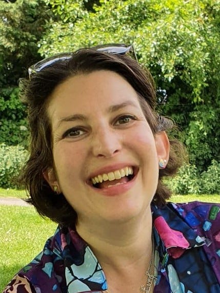 A white woman with short dark hair, standing outside in front of a tree and laughing (with sunglasses on her head)