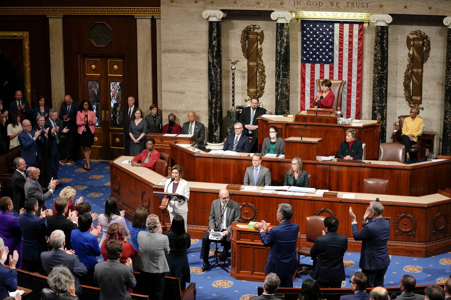 Nancy Pelosi, in a white pant suit, speaks on the House floor. She is receiving a standing ovation from her colleagues.