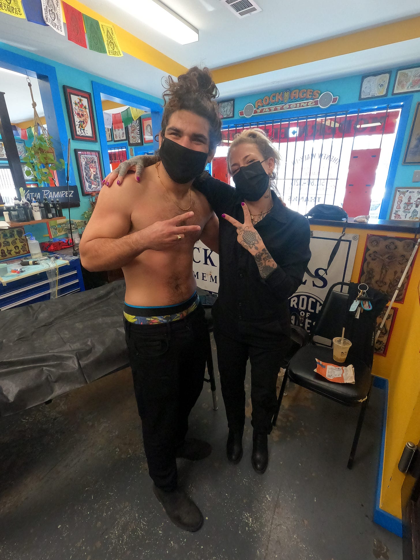 anthony and the tattoo artist hold up peace signs with their hands. Anthony is smiling and has no shirt on