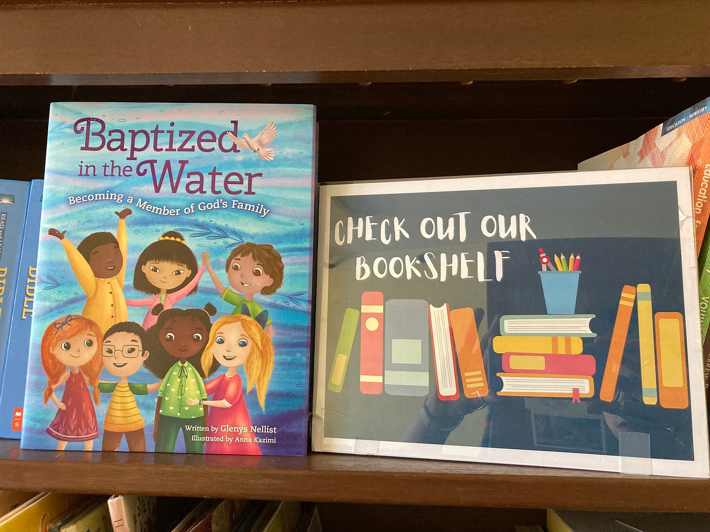 A copy of the children's book Baptized in the Water stands on a shelf next to a sign reading Check Our Our Bookshelf.