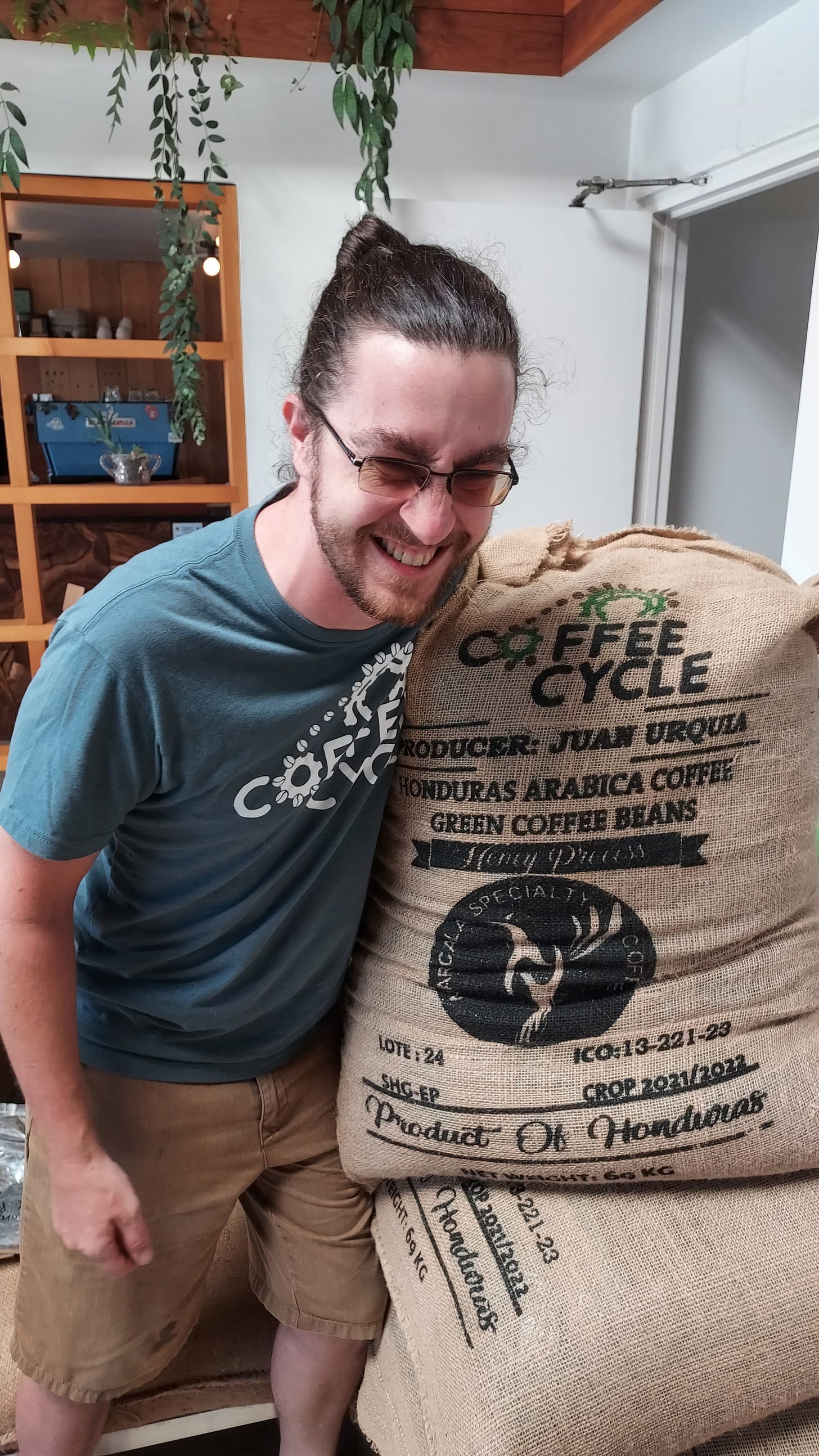 A tall white man with a scruffy beard, hair pulled back, glasses, blue t-shirt has his arm around a burlap coffee sack and is doubled over with a big smile on his face
